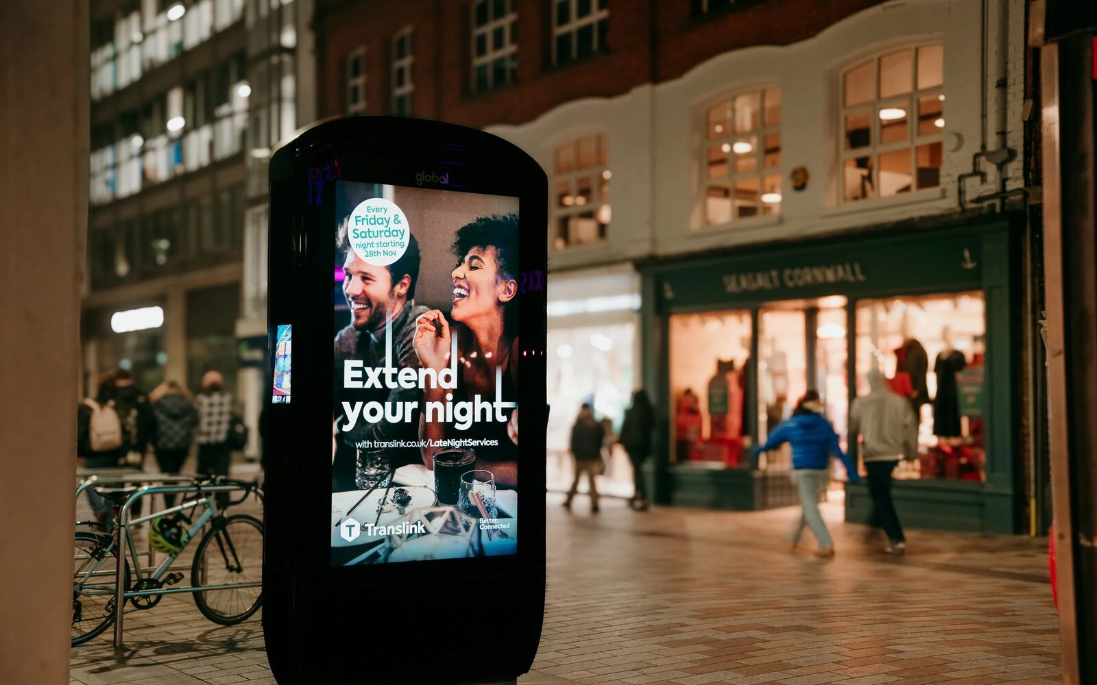 A Pulse smart hub standing on a city high street at dusk, its screen lit with an advertisement as shoppers pass